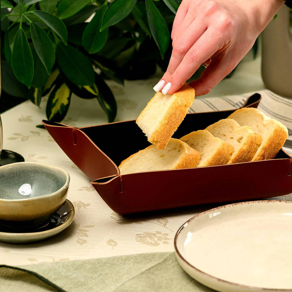 Modern restaurant table setting featuring a minimalist brown leather bread basket. The photo captures a hand picking up a slice of fresh baguette from the handcrafted leather valet tray, showcasing a sophisticated way to serve appetizers in a contemporary dining space