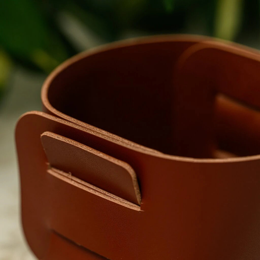 A close-up of a small, square bread bin made from stiff brown leather. The design features a unique interlocking system where leather tabs are inserted into slots to hold the shape without stitching. It sits on a patterned tablecloth with a blurred plate in the foreground