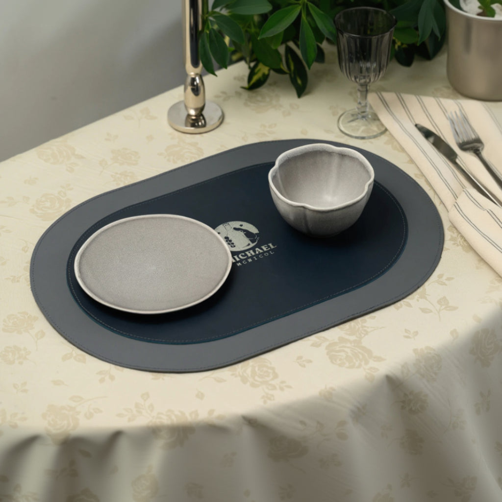 A top-down view of a dual-textured oval leather placemat serving as the centerpiece of a formal table setting. The mat has a smooth outer border stitched to a matte inner section with an embossed logo for "MICHAEL MCNICOL" in the center. A ceramic bowl and plate are arranged on the mat, and the table is set with a floral tablecloth, a striped napkin, and glassware.