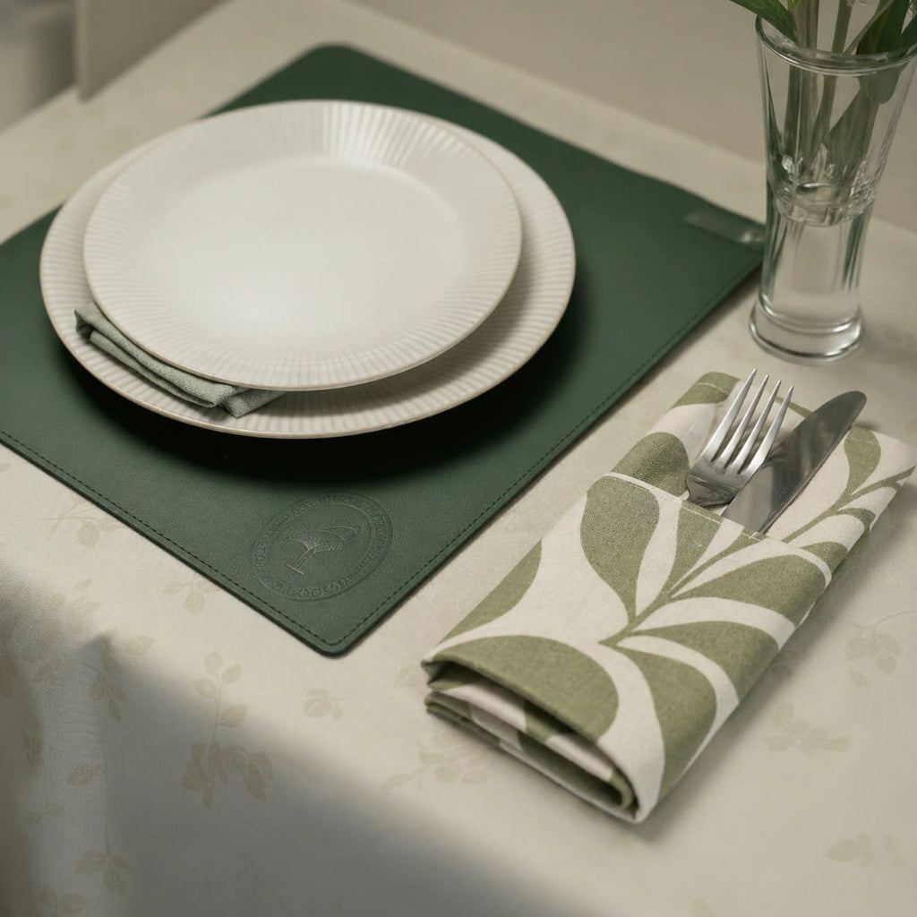 A close-up view of a square forest green leather placemat featuring a debossed circular logo in the corner, styled with a ribbed white plate and a green and white leaf-patterned napkin holding silver cutlery.