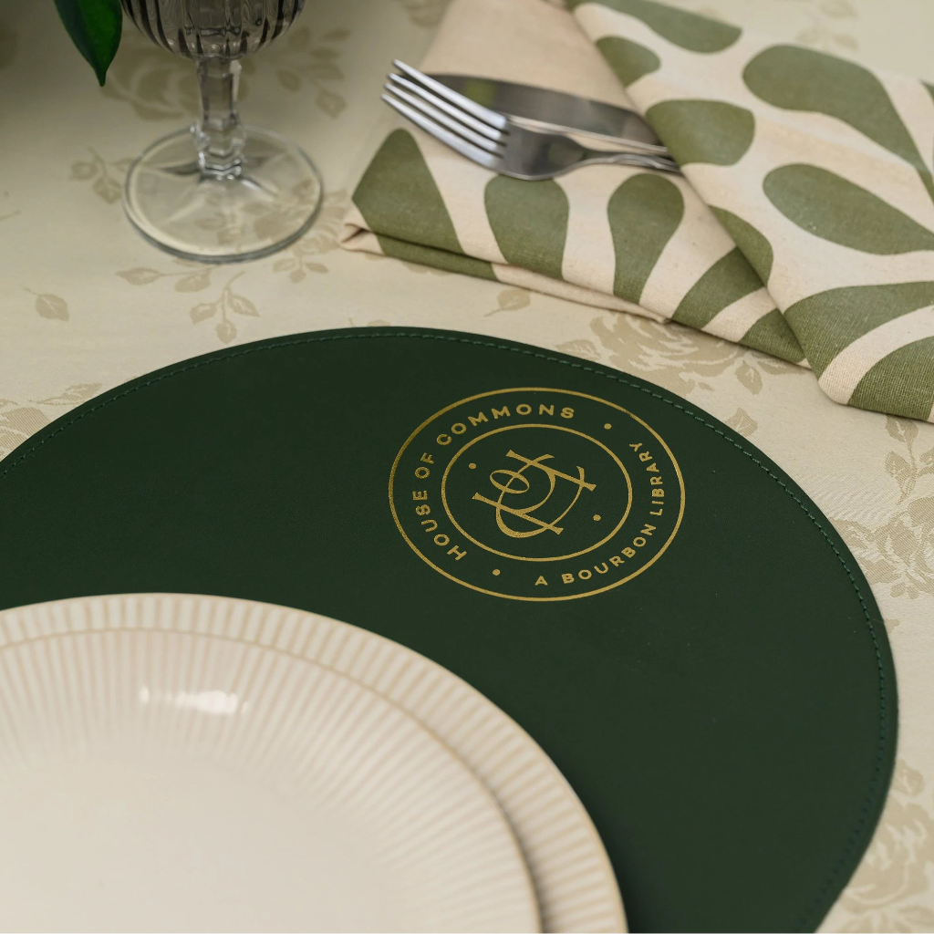 A detailed view of a forest green leather round placemat featuring a central gold foil embossed seal that reads 'HOUSE OF COMMONS A BOURBON LIBRARY' with a custom monogram. The placemat is on a beige damask tablecloth with a ridged plate, green-patterned napkins, and silver cutlery