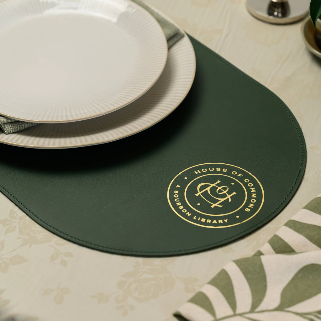 Oval forest green leather placemat with a gold foil embossed logo for "House of Commons A Bourbon Library." The image shows a top-down view of the mat styled with a white ribbed ceramic plate, a green linen napkin, and detailed perimeter stitching on a light floral tablecloth