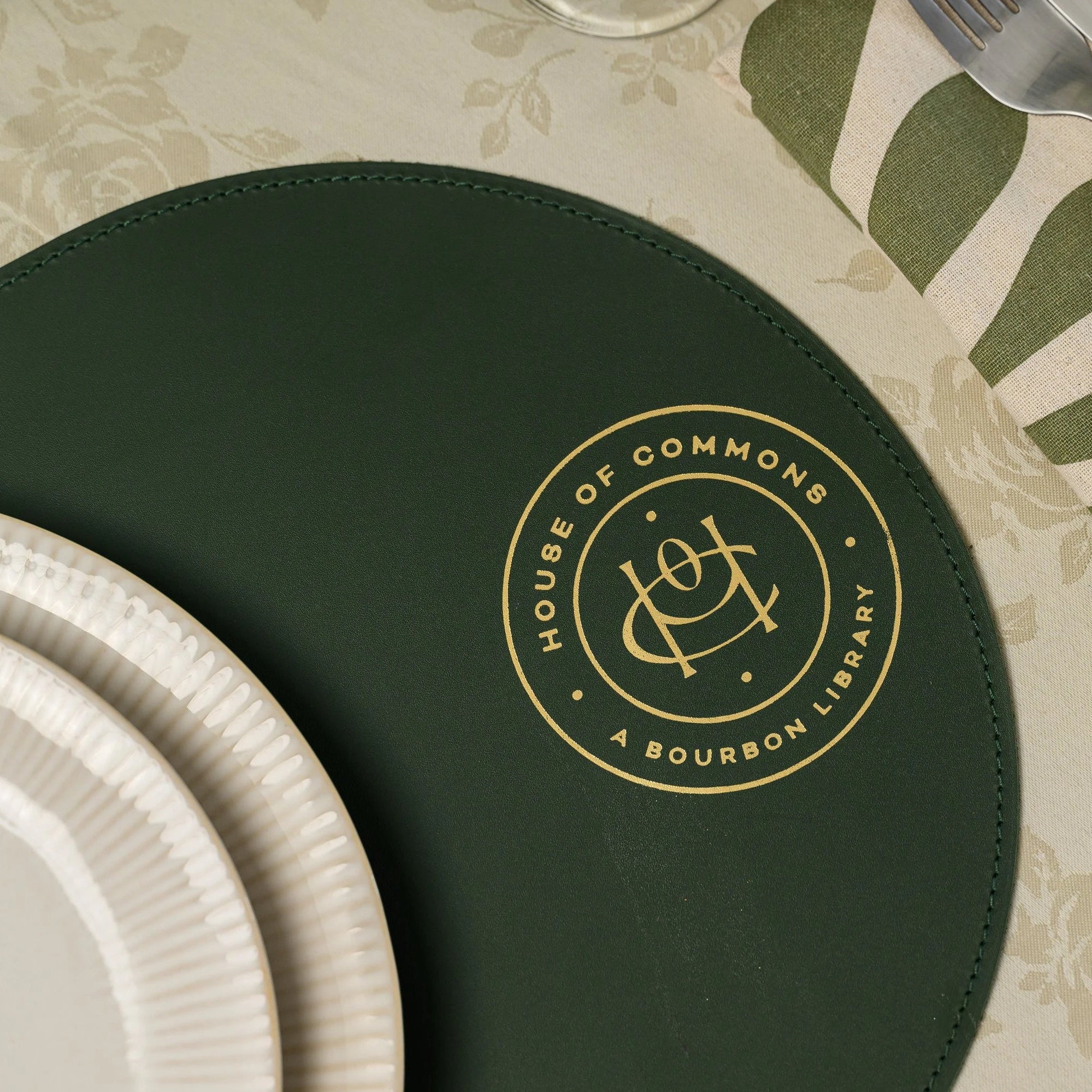 Top-down view of a premium dark green leather round placemat. The design features a detailed, reflective gold metallic embossed seal for 'House of Commons A Bourbon Library'. Shown with stacked ceramic dishes on a beige damask tablecloth.