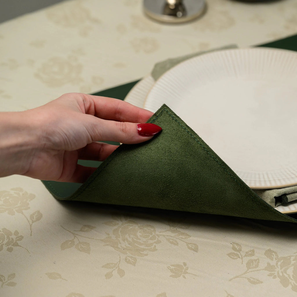 A close-up, high-angle shot of a premium forest green leather placemat featuring an elegant gold foil embossed logo of "House of Commons A Bourbon Library." The image captures the rich texture of the leather and the reflective metallic gold seal, set against a cream floral tablecloth with a white ribbed plate.