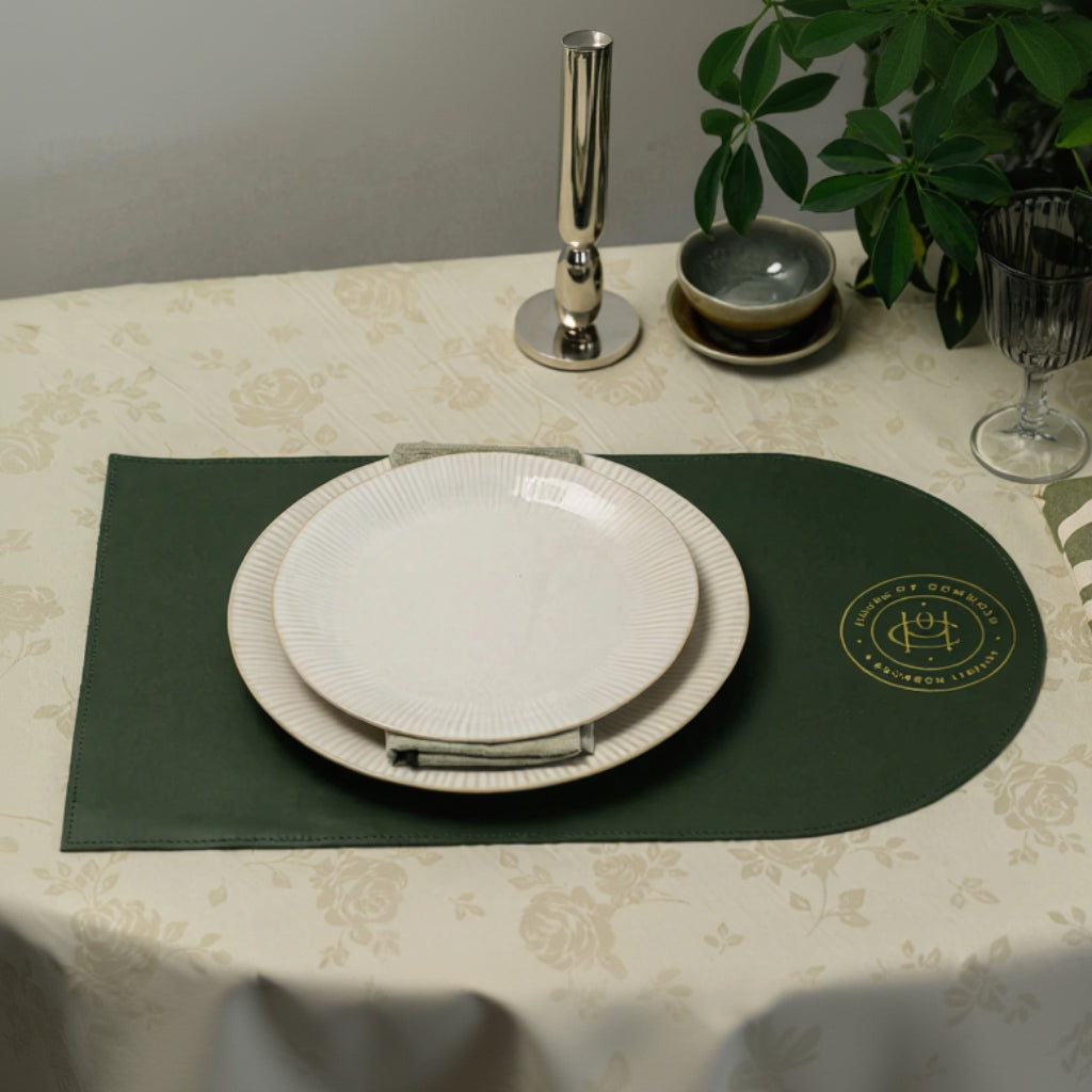 Top-down lifestyle view of a forest green D-shaped leather placemat on a cream floral tablecloth. The mat features a gold foil embossed "HOUSE OF COMMONS A BOURBON LIBRARY" logo and is styled with stacked white ribbed plates, a silver candlestick, and green foliage in the background