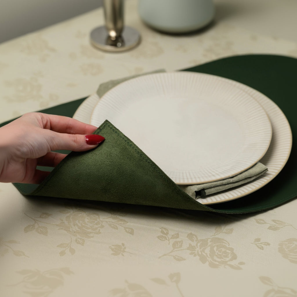 A hand with red manicure lifts the corner of a dark green leather placemat, showing its soft suede texture. A cream-colored ribbed plate and a linen napkin are placed on top.