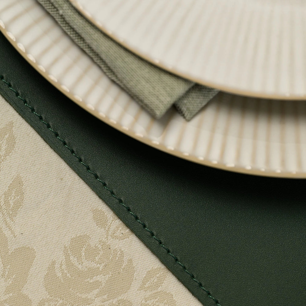 Extreme close-up of a dark green leather placemat showing decorative perimeter stitching. A ribbed ceramic plate and a sage green linen napkin rest on its smooth surface.