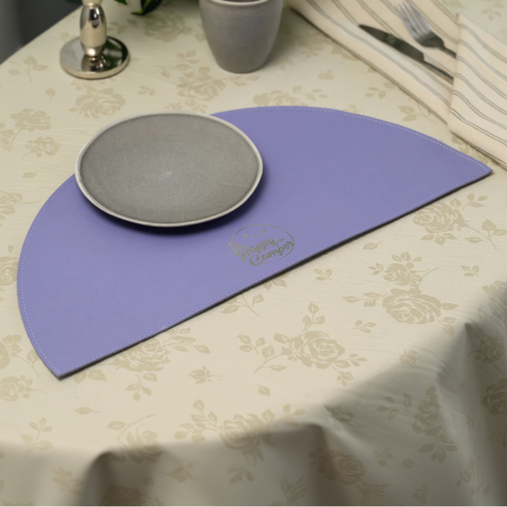 A top-down lifestyle shot of a semi-circular lavender leather placemat on a cream floral tablecloth, featuring a grey ceramic plate and a silver-engraved "The Happy Camper" logo.