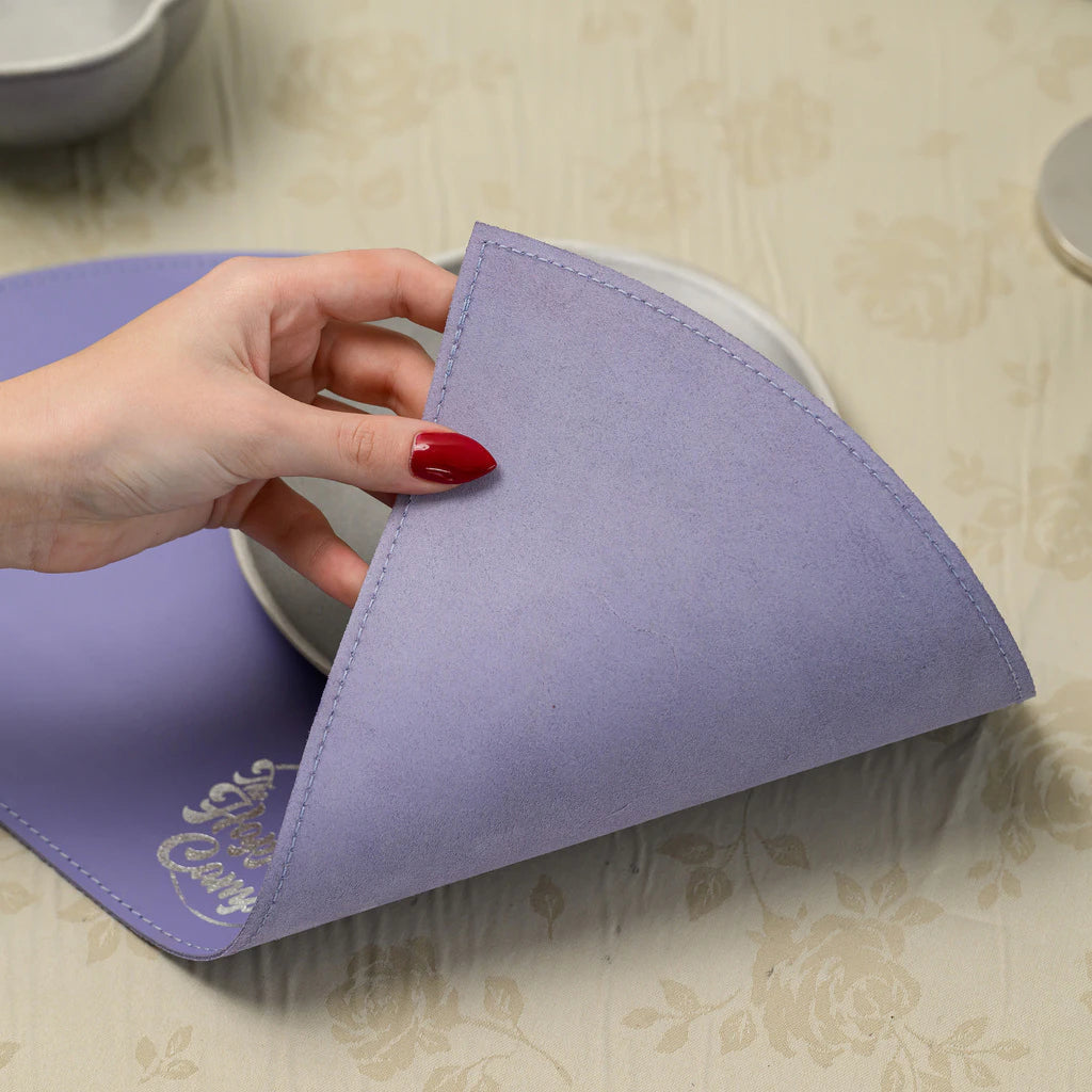 A hand with red nails peeling back the corner of the lavender placemat to demonstrate its flexibility and the high-quality texture of the material.