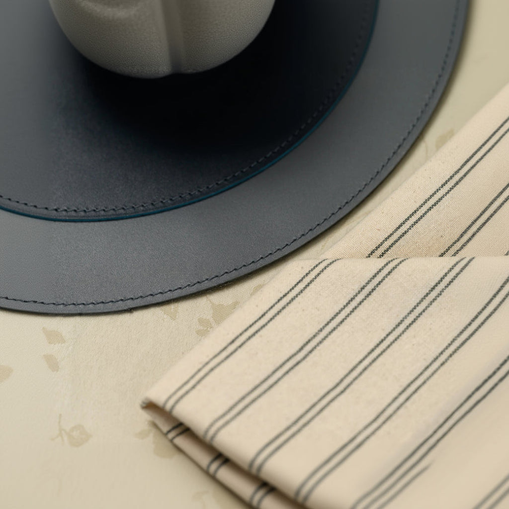 A close-up view focusing on a leather placemat with two distinct textures and visible perimeter stitching. The smooth outer border is seamlessly stitched to a matte inner section. Part of a striped fabric napkin and the base of a ceramic bowl are visible, with all items resting on a patterned tablecloth.