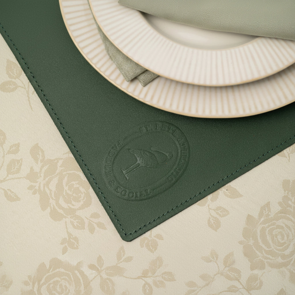 Detailed shot showing the fine texture and quality stitching of a square dark green leather table mat. A detailed circular debossed brand seal is prominent in the corner, resting on a damask floral tablecloth