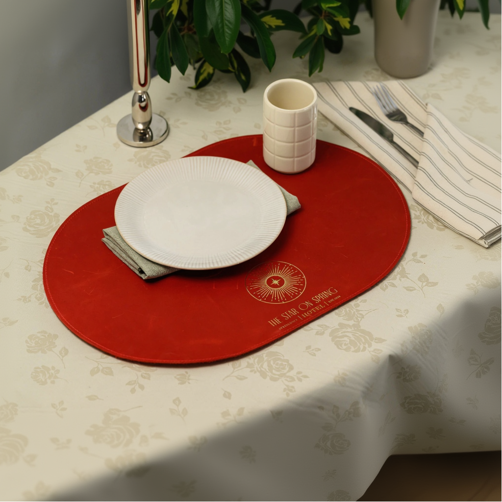 Luxury red oval leather placemat with decorative gold lettering, presented in a sophisticated table setting for hospitality or upscale home decor