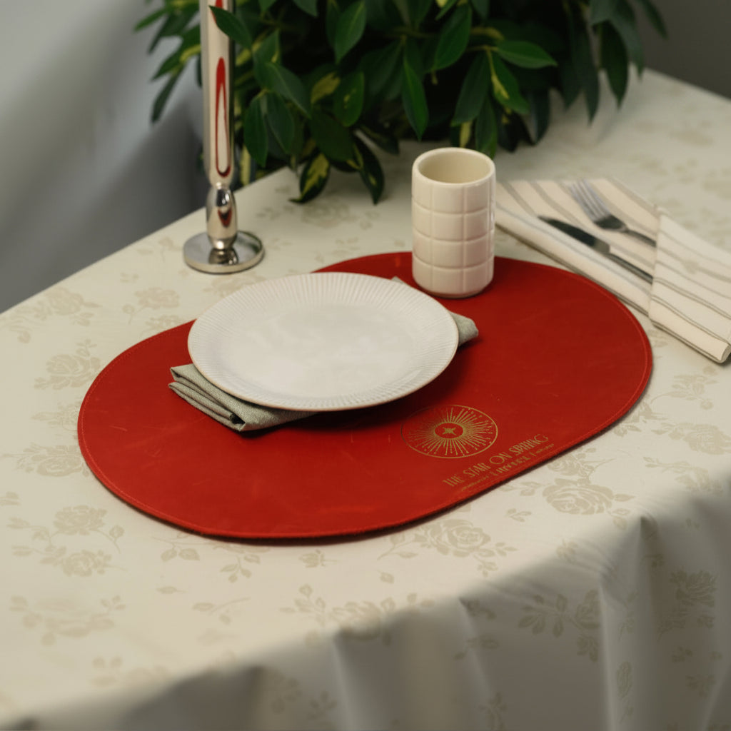 Top-view of a luxury oval red leather placemat with a gold foil hotel logo, featuring white ribbed plates, a ceramic cup, and striped napkins on a cream floral tablecloth