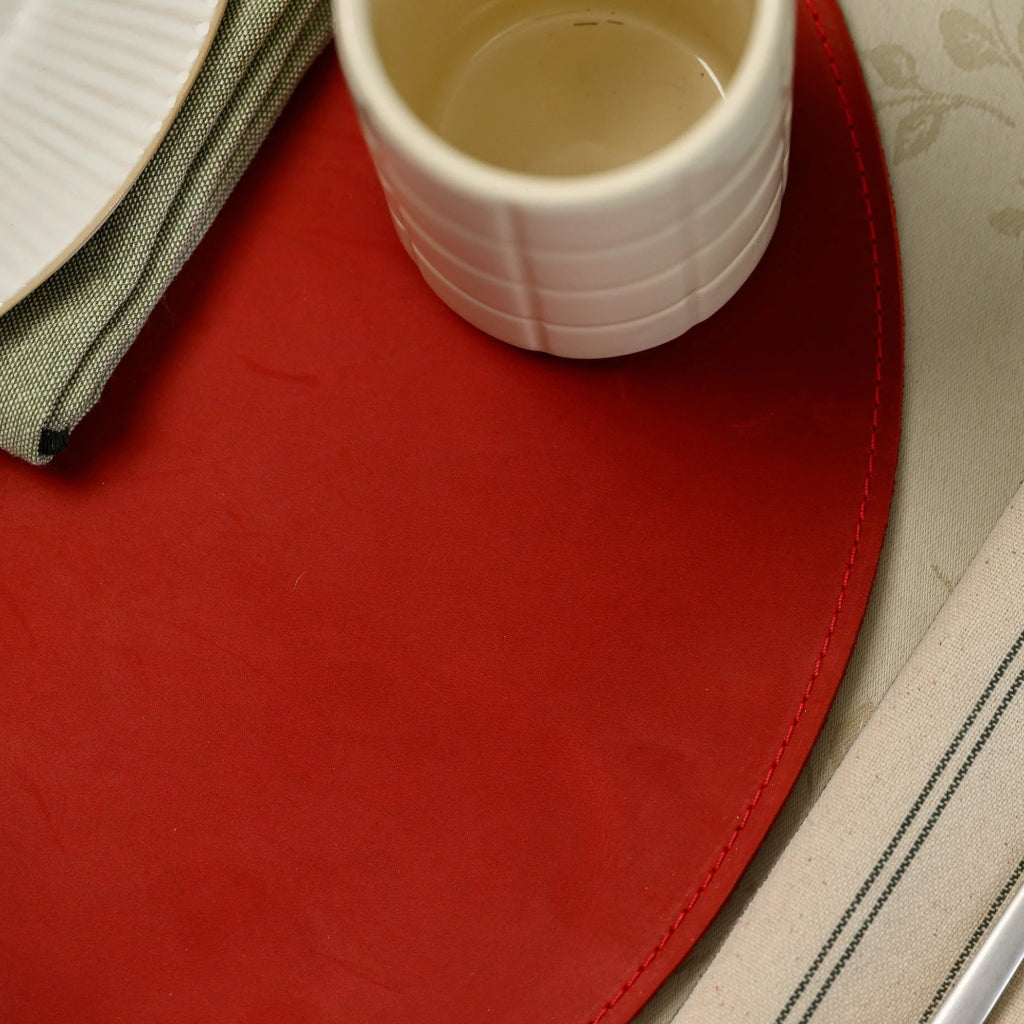 Macro view of an oval red leather placemat showing professional edge stitching, a cream ceramic cup, and a white ribbed plate on a floral tablecloth