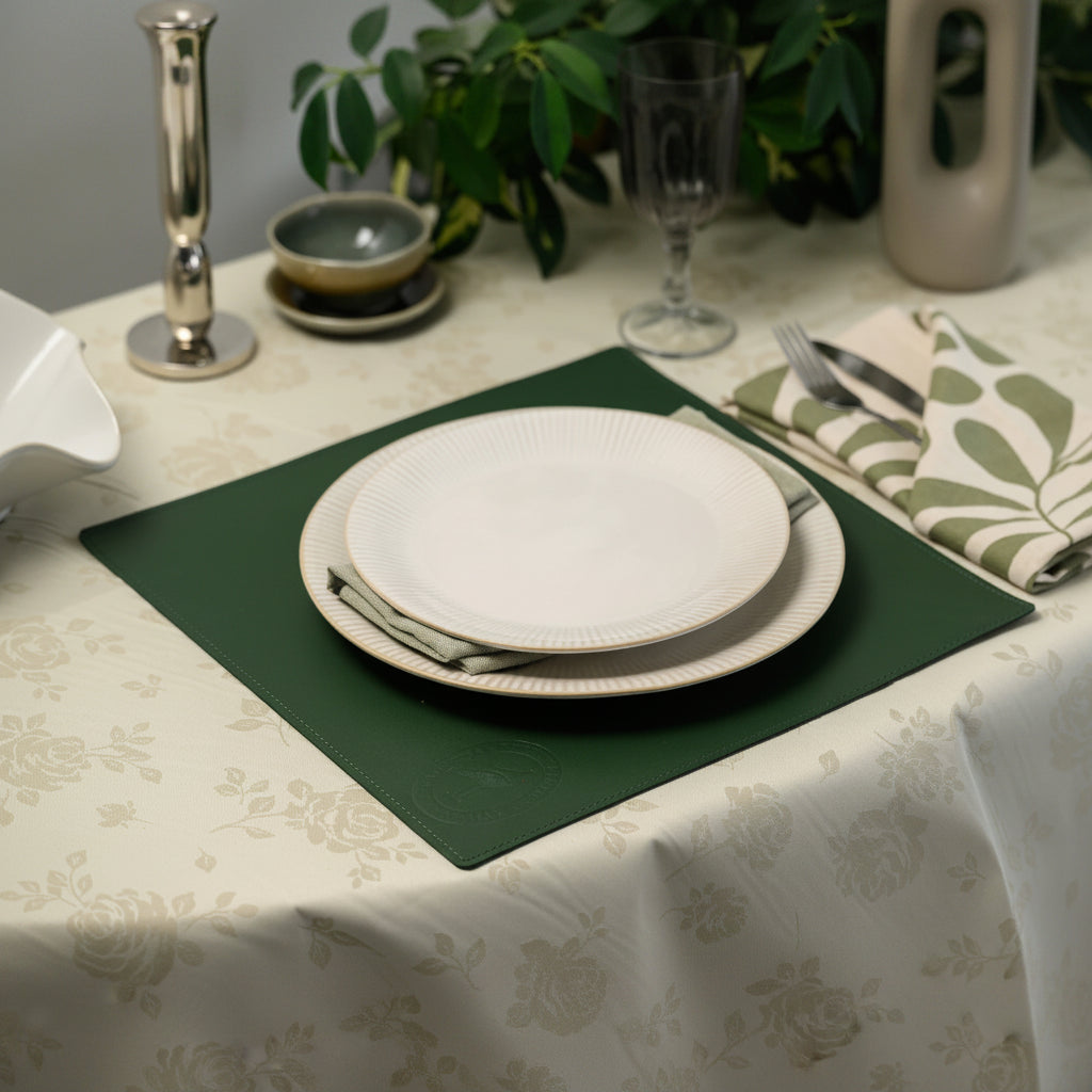 Elegant forest green leather placemat with stitched edges and embossed logo, styled on a white floral tablecloth with ribbed plates, cutlery, and sage green botanical napkins