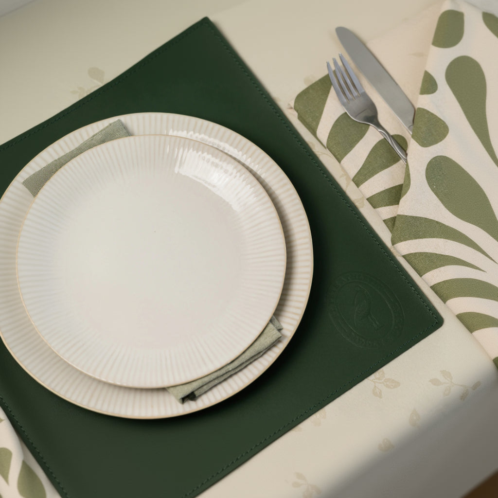 Top-down view of a square forest green leather placemat with an embossed brand logo, featuring stacked white ribbed ceramic plates and elegant silver cutlery on a botanical-patterned napkin