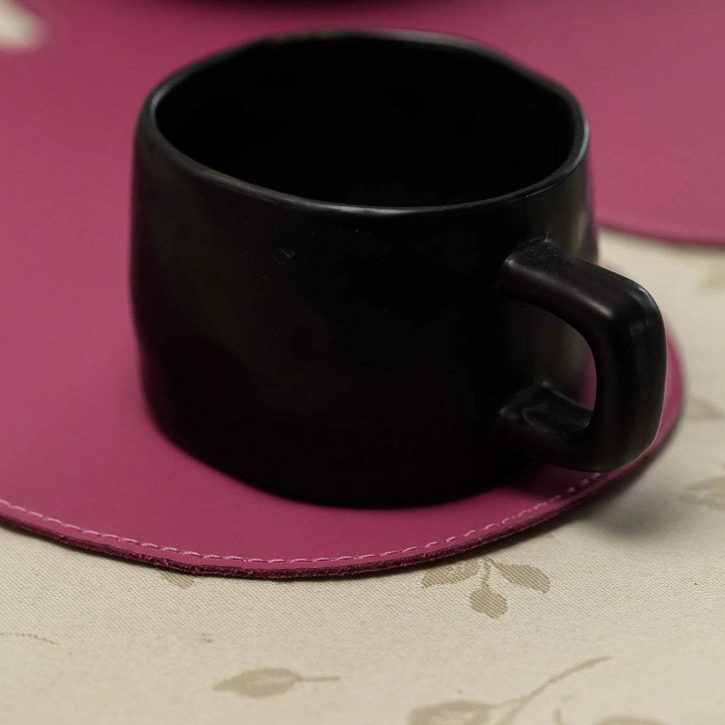 A close-up shot of a modern, matte black mug with a unique, slightly asymmetrical shape. It rests on a circular magenta or deep pink leather coaster with visible stitching around the edge, placed on a light-colored patterned tablecloth.