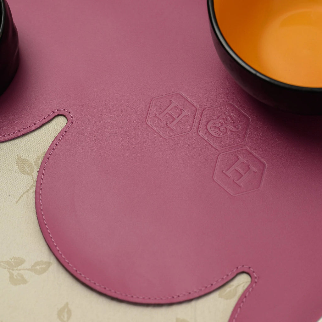 A close-up of a magenta leather placemat with a unique wavy edge and decorative stitching. The surface features an embossed logo consisting of three hexagonal shapes with letters.