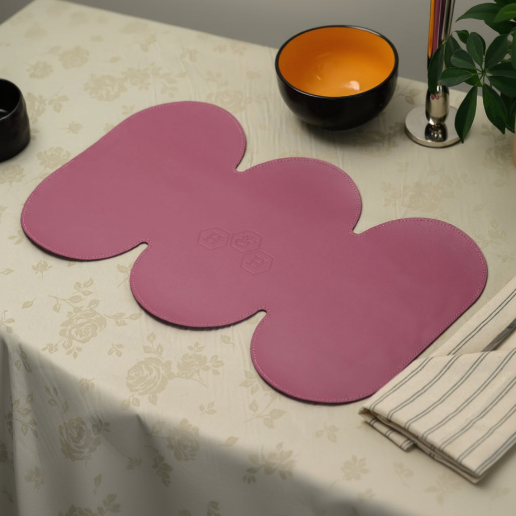 A large pink leather placemat with a unique triple-lobed cloud shape and stitched edges. The placemat features a central embossed hexagonal logo and is laid out on a cream floral tablecloth.