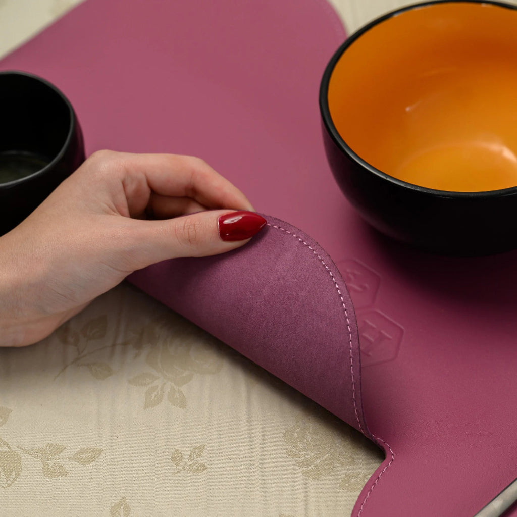 A hand with red nails lifts the edge of a magenta leather placemat to reveal its soft, purple suede underside. The placemat has a unique wavy shape and neat perimeter stitching.