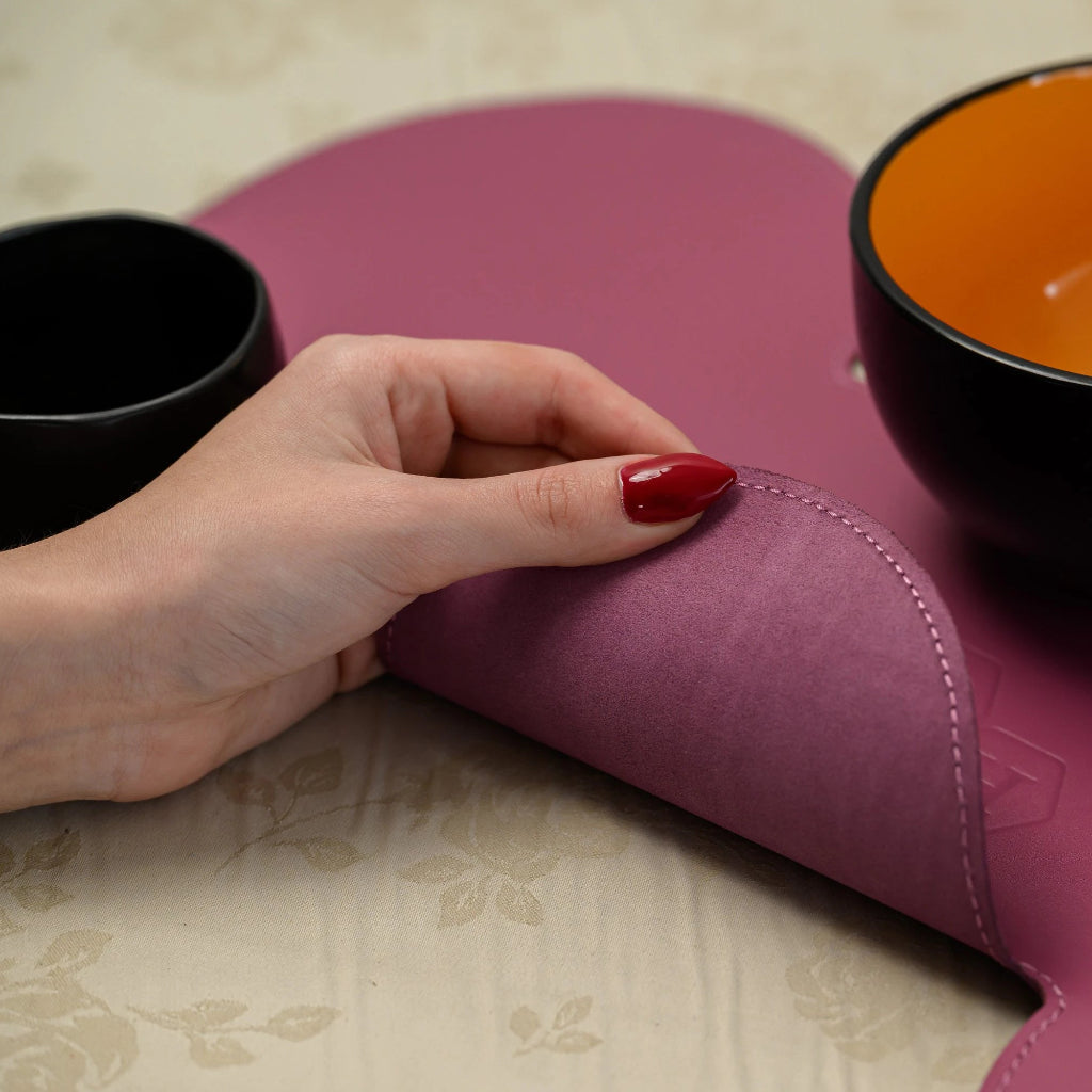 Close-up of a person lifting the corner of a cloud-shaped fuchsia placemat, highlighting the contrast between the smooth leather top and the textured suede reverse side. A modern black ceramic bowl with an orange interior sits on the surface.