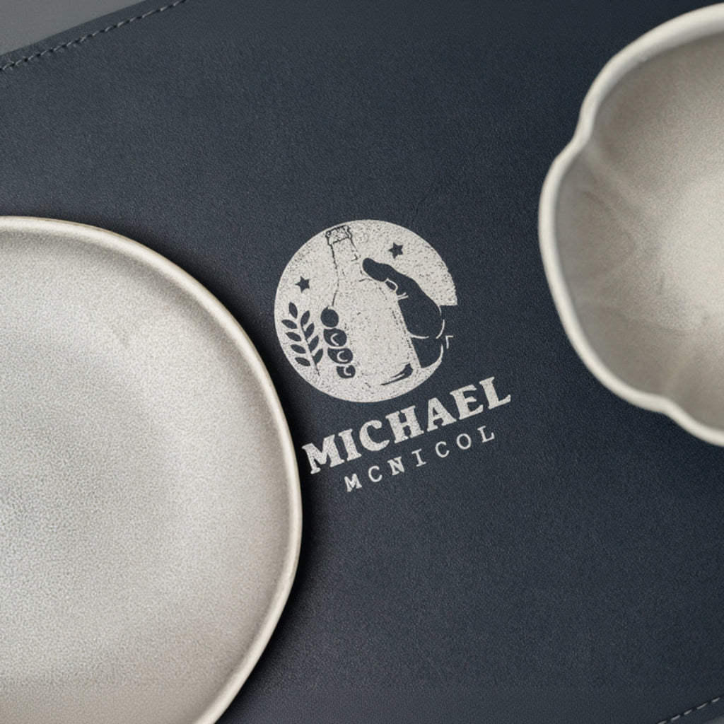 An overhead close-up of a leather-textured placemat featuring a detailed white logo. The logo depicts a fist clutching a bottle, a sprig of barley, and stars inside a circle. The name 'MICHAEL MCNICOL' is printed below. Flanking the logo are parts of two light grey, irregular-shaped ceramic dishes.