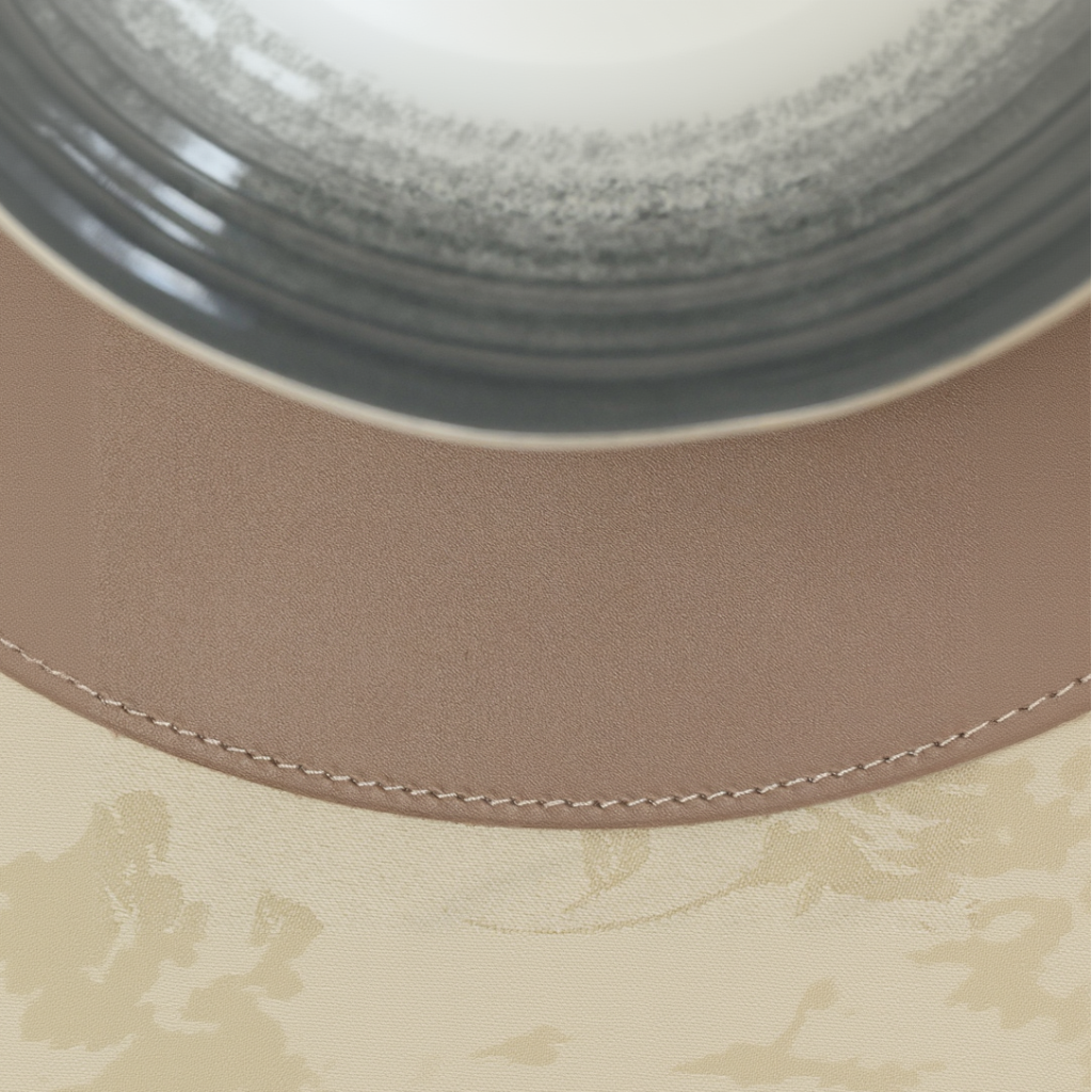 A macro top-down shot of the leather placemat, emphasizing the precise perimeter stitching along its curved edge. The smooth, matte texture of the leather is shown in detail, positioned between a ceramic bowl and a floral patterned tablecloth. The lighting highlights the fine detail of the thread and the premium finish of the material