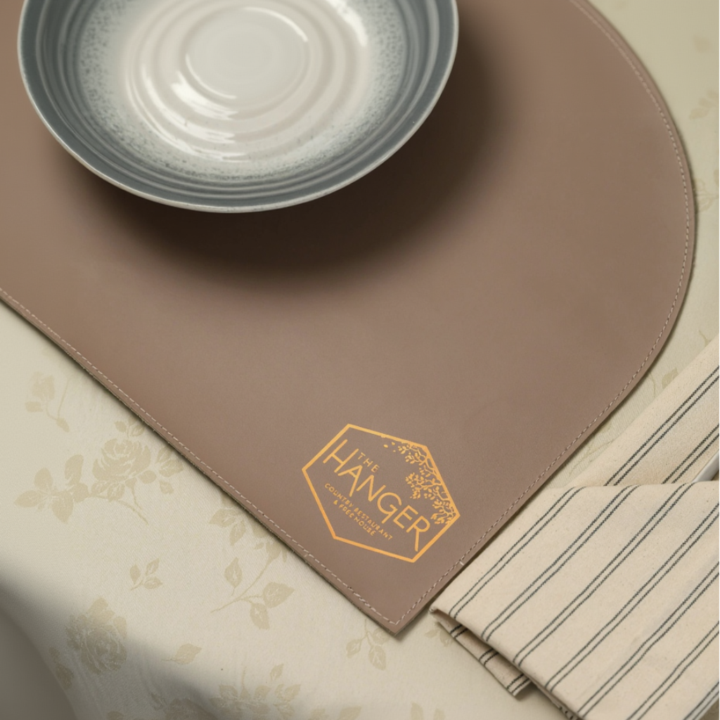 A top-down view of a rectangular leather placemat with two rounded diagonal corners and detailed perimeter stitching. A metallic hexagonal logo for "The Hanger" is embossed in one corner. The placemat is part of a refined table setting, featuring a ceramic plate, a glass, and a striped fabric napkin with cutlery, all placed on a floral-patterned tablecloth.