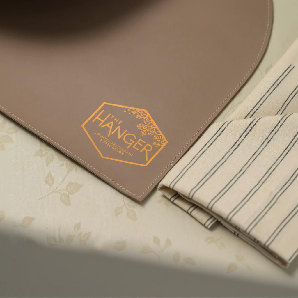A close-up, angled shot of a leather placemat with two rounded diagonal corners and neat perimeter stitching. The surface features a metallic hexagonal logo for "The Hanger Country Restaurant & Free House" decorated with a delicate leaf motif. The placemat is set on a floral-patterned tablecloth next to a folded, striped fabric napkin.