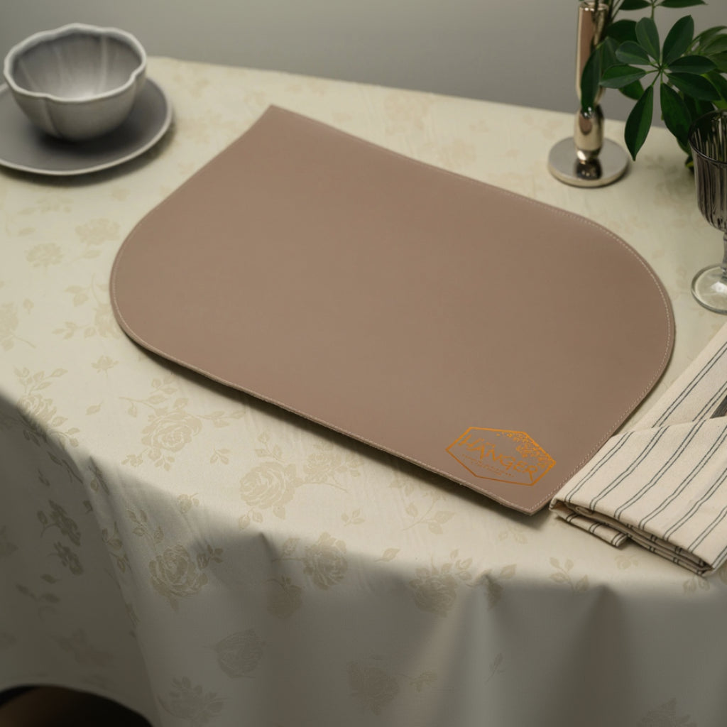 A top-angle view of a mocha-colored leather placemat with a unique asymmetrical semi-oval shape. The placemat features a smooth matte surface with a subtle orange logo embossed in the bottom corner and fine stitching along the edges. It is laid out on a cream floral tablecloth next to a striped napkin with cutlery, a wine glass, and a small bowl, showcasing its modern and minimalist design
