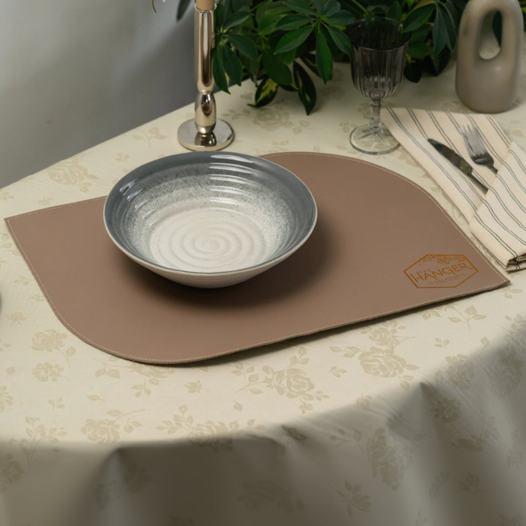 A wide-angle view of a professionally set table featuring a leather placemat with two rounded diagonal corners and fine perimeter stitching. A bowl is centered on the placemat, which is embossed with a metallic hexagonal logo. The table is decorated with a floral tablecloth, a striped napkin with silverware, glassware, a candlestick, and a decorative vase with green foliage in the background.