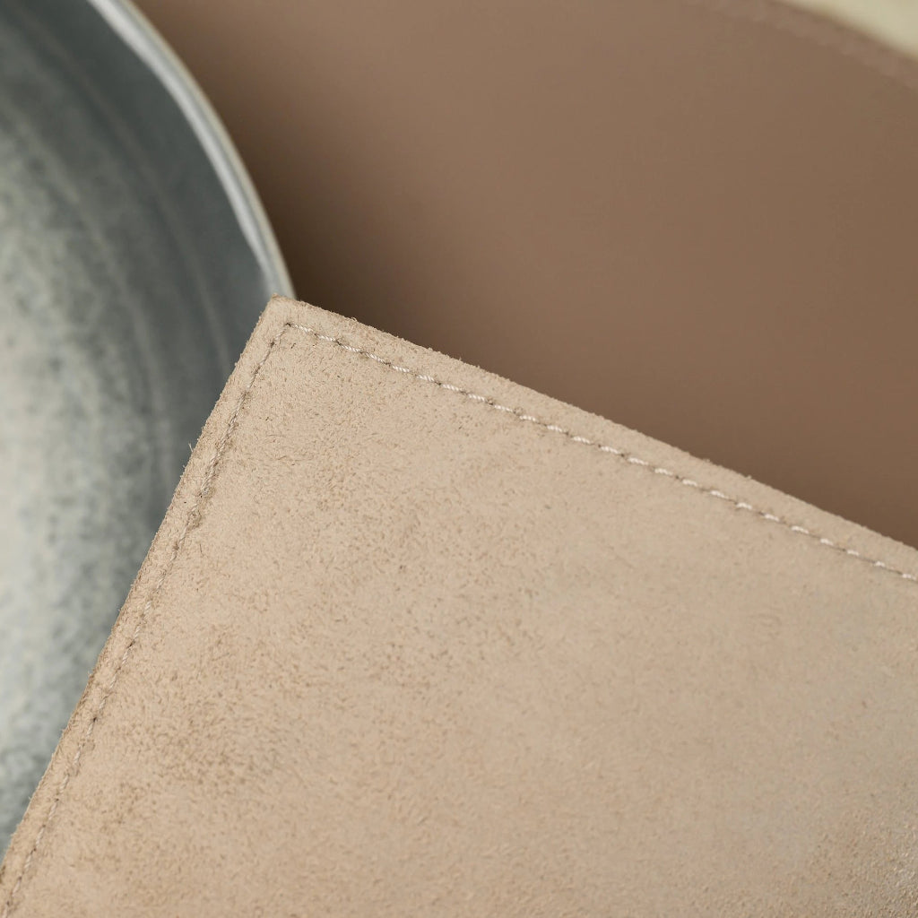 An extreme close-up focusing on the corner of a leather placemat, showing the fibrous, napped texture of its underside. The image highlights the clean, precise perimeter stitching along the straight edge. In the blurred background, the smooth top surface of the placemat and a portion of a ceramic bowl are visible.