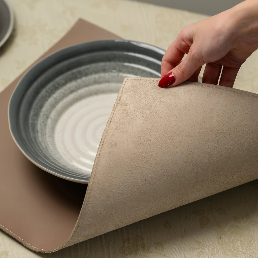 A person's hand lifts the corner of a leather placemat to reveal the natural, fibrous texture of its underside. The shot demonstrates the flexibility and thickness of the material, as well as the consistent perimeter stitching. The placemat is part of a table setting with a ceramic bowl on top, resting on a floral tablecloth.