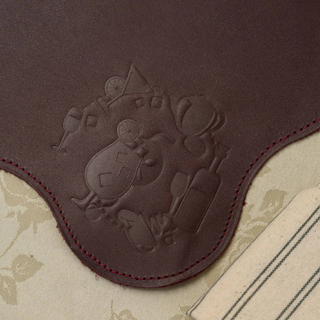 A close-up view of the detailed embossing on a leather placemat with a wavy, scalloped edge. The design features various cocktail glasses, an orange slice, and a bottle. The mat is finished with precise perimeter stitching and is set on a patterned tablecloth.