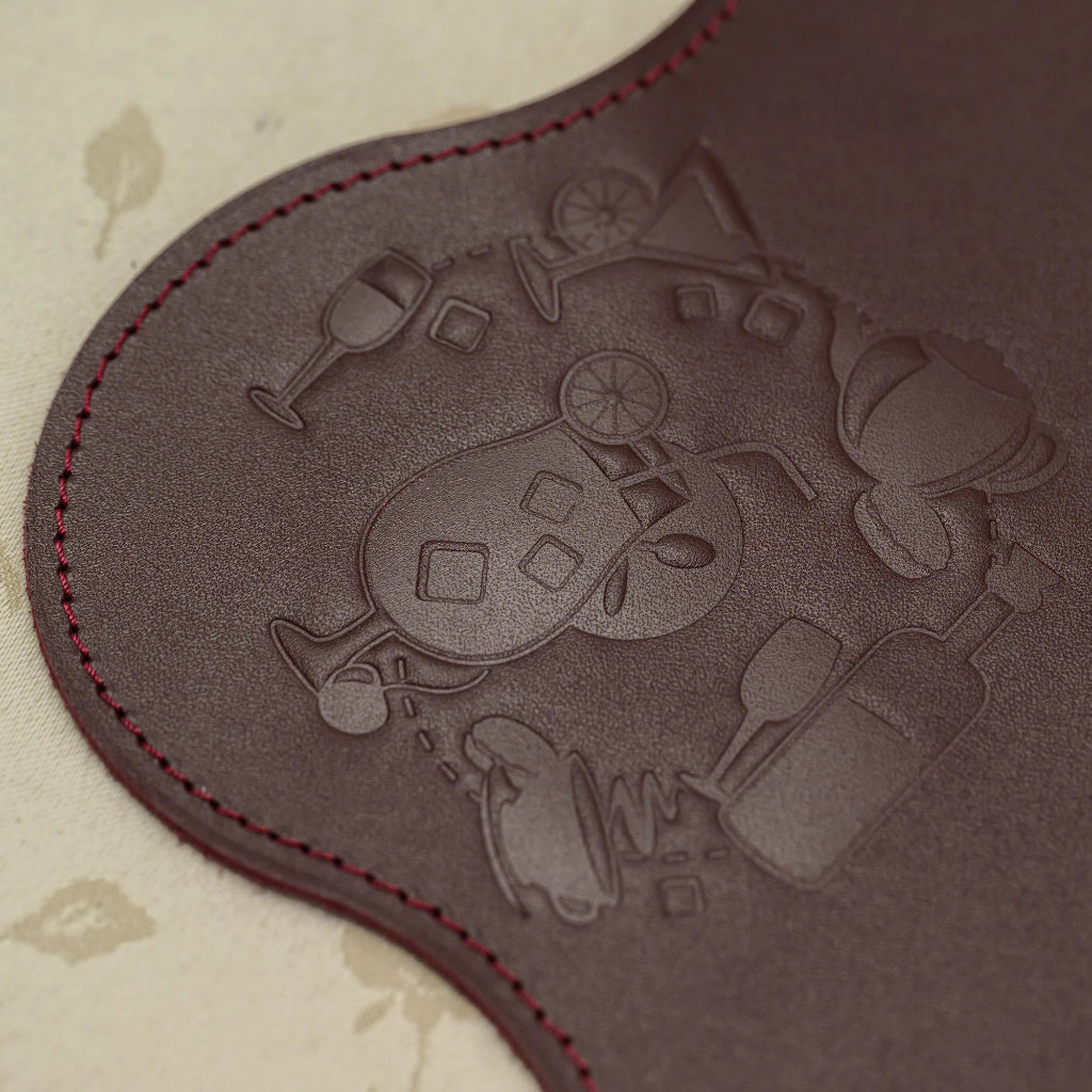 Close-up of a premium burgundy leather product featuring a debossed circular design of various beverage icons including cocktails, wine, coffee, and tea. The image shows high-quality red perimeter stitching and a smooth leather grain texture against a neutral floral background.
