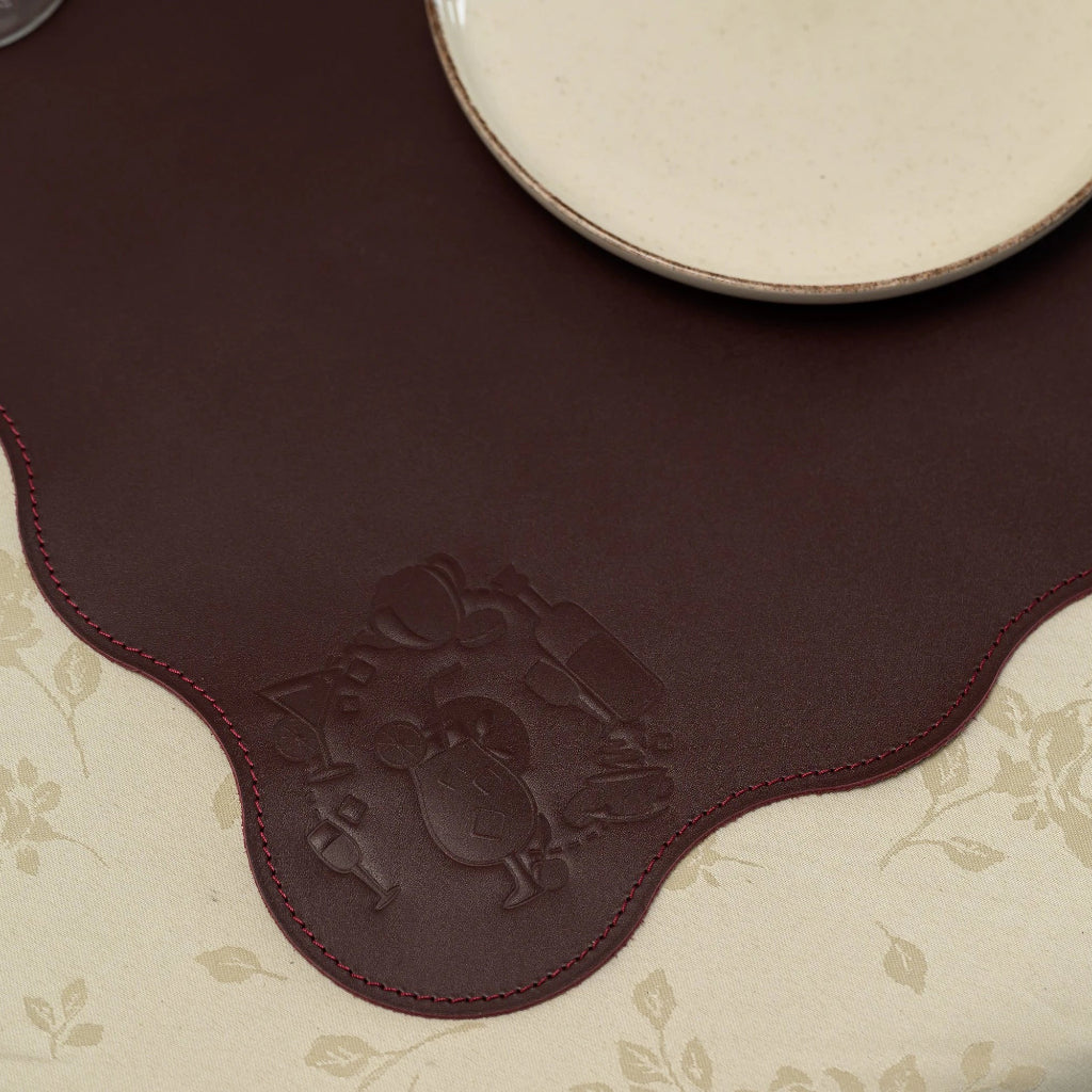 A close-up, top-down view of a leather placemat with a unique wavy, scalloped edge. The mat features a detailed embossed illustration in the corner, depicting various cocktail glasses and a bottle. It is finished with tonal perimeter stitching and is shown as part of a table setting on a floral-patterned tablecloth.