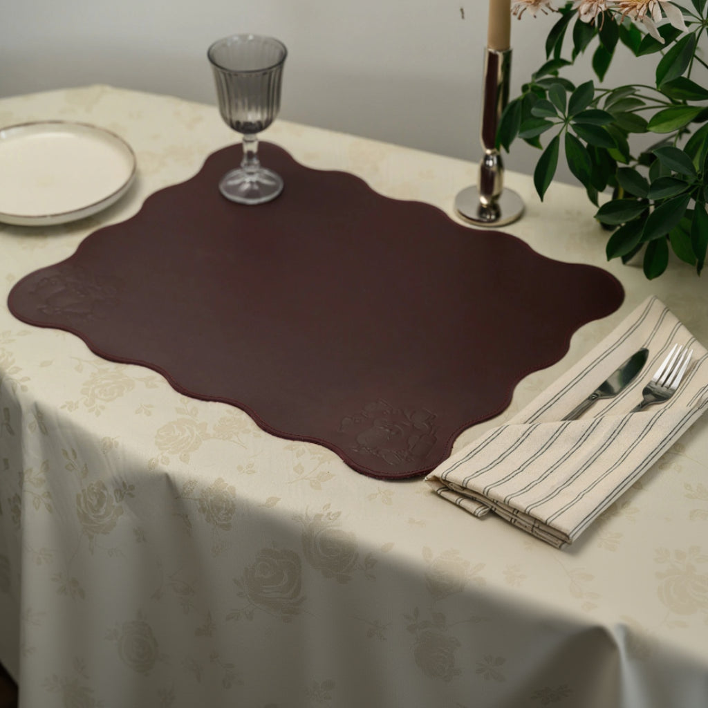Large scalloped-edge burgundy leather placemat on a cream floral tablecloth. The mat features wavy borders and debossed beverage-themed illustrations in the corners. Styled with a glass, silver candlestick, and striped linen napkin with cutlery