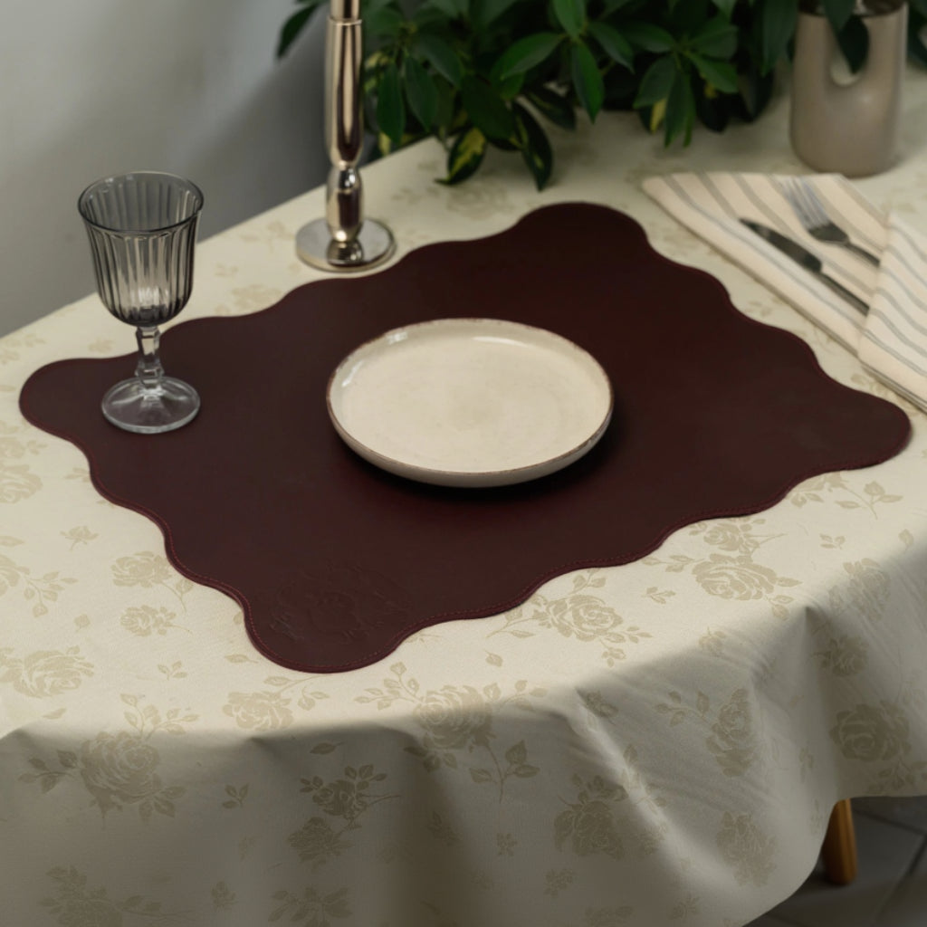 Dark burgundy leather placemat with a large scalloped edge design. It features subtle, debossed decorative icons and sits on a cream floral-patterned tablecloth. Styled with a white plate, grey stemware, striped linen napkins, and silver cutlery in a sophisticated table setting.