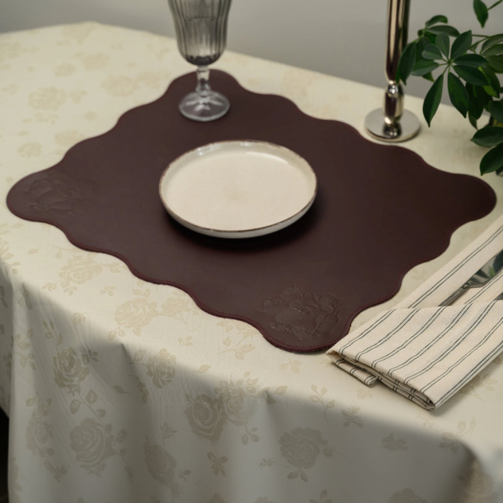 Large burgundy leather placemat with a unique scalloped wavy edge on a cream floral damask tablecloth. The mat features a subtle debossed corner design and is styled with a white ceramic plate, a grey glass, a silver candle holder, and a striped linen napkin
