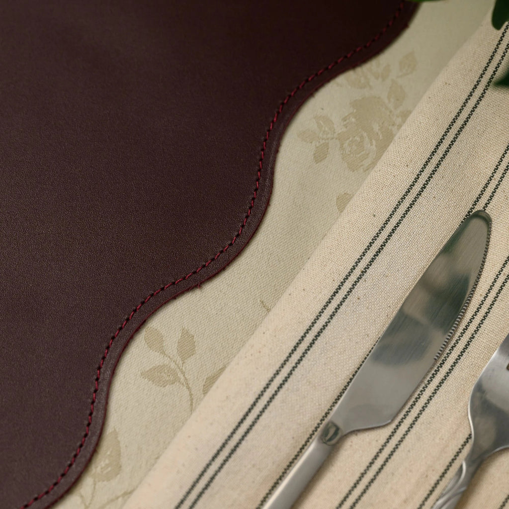 Extreme close-up of a burgundy leather placemat showing the fine grain texture and professional red perimeter stitching along a scalloped wavy edge. A striped linen napkin with a silver knife is placed alongside on a cream floral tablecloth