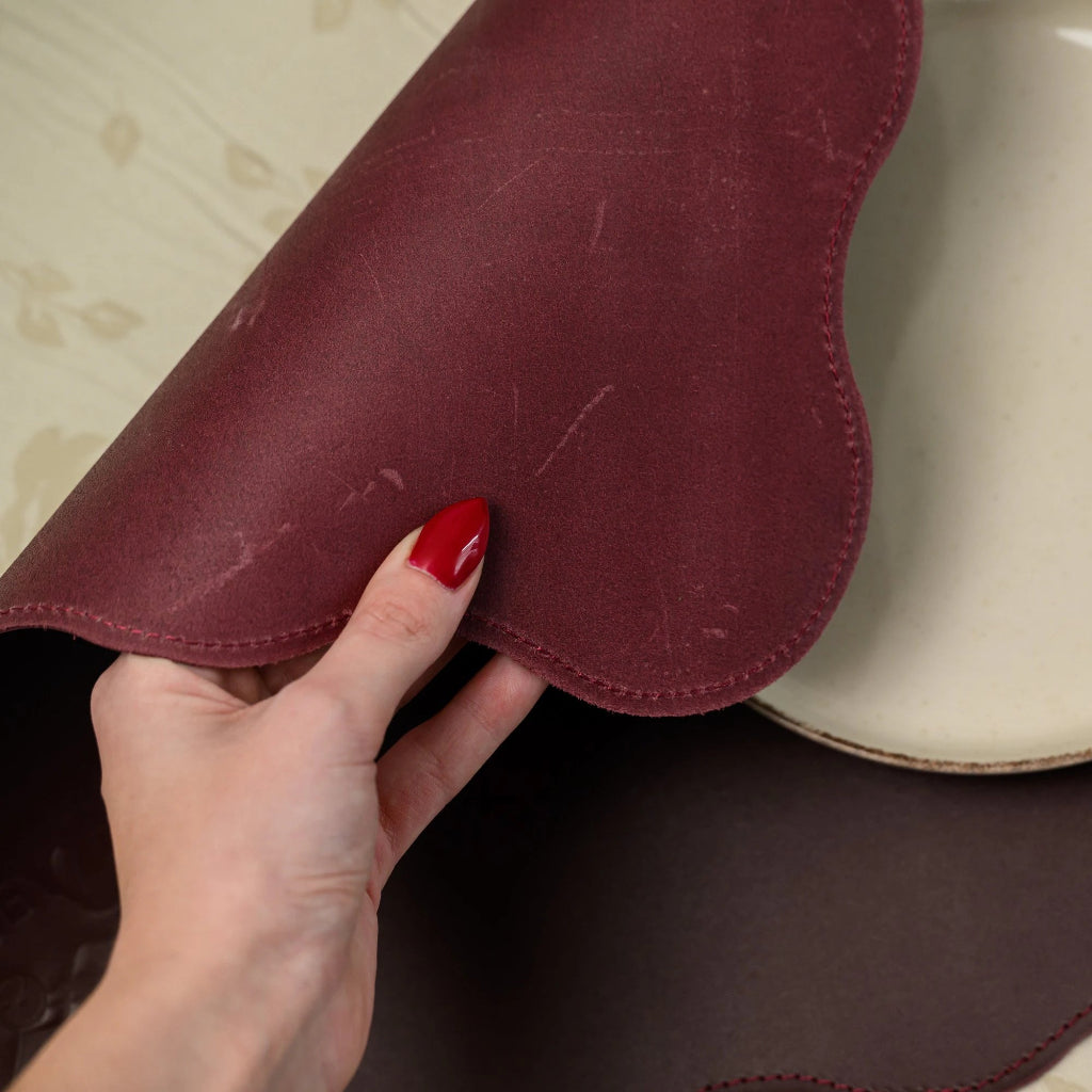 A hand with red nails lifting the scalloped edge of a burgundy leather placemat to show its flexible material. The image features a top-down view of the matte leather texture, professional red stitching, and a cream-colored ceramic plate on a floral tablecloth