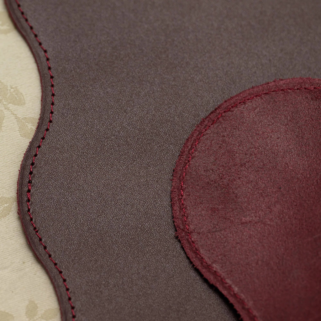 Close-up of a burgundy leather placemat featuring a debossed circular design of beverage icons, including cocktails, wine, coffee, and tea. The image highlights the smooth leather grain and professional red perimeter stitching.