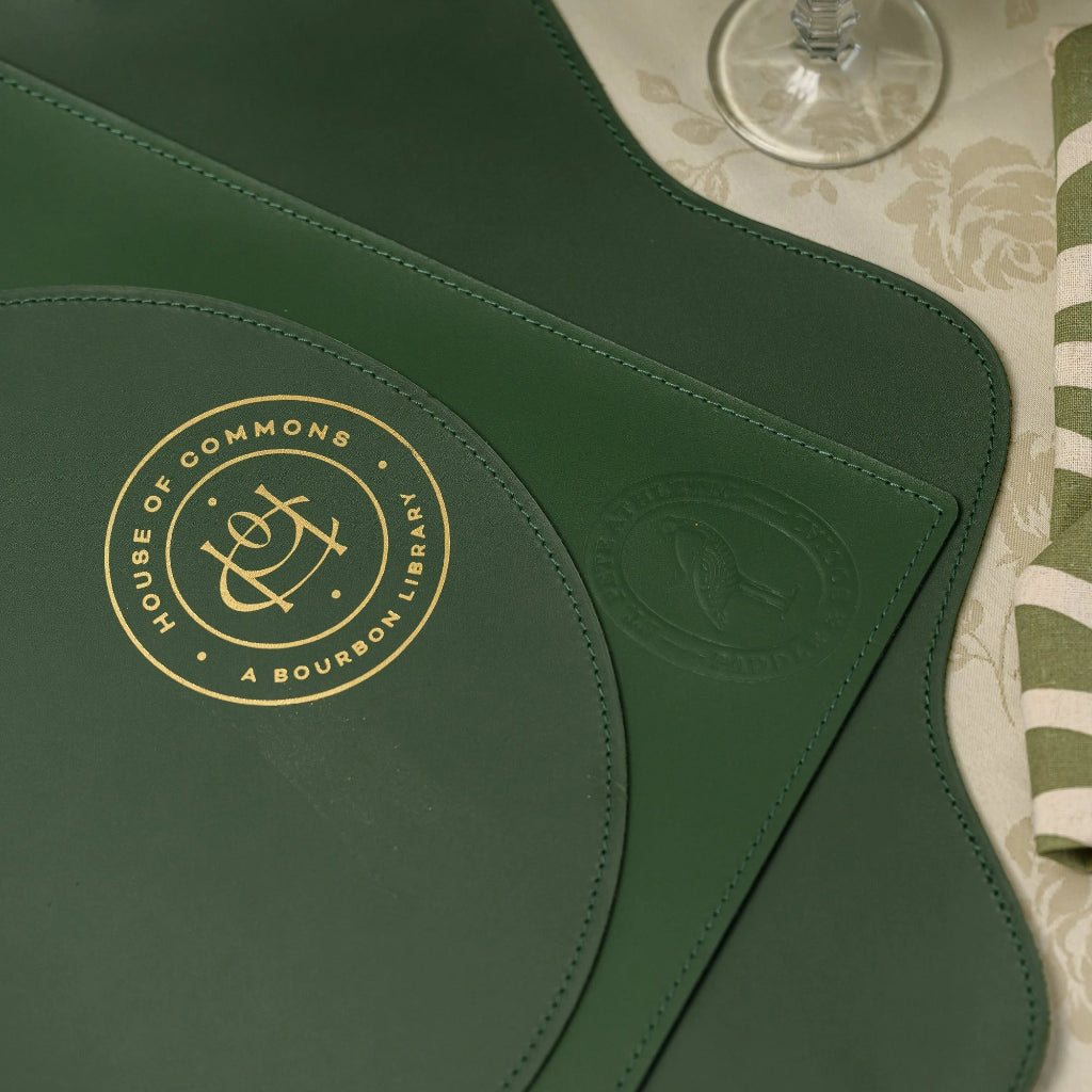 ІDetailed shot showing the variety of green leather table mats, including a circular-edged pad with ornate gold lettering, paired with botanical print napkins and silver cutlery for an upscale dining ambiance