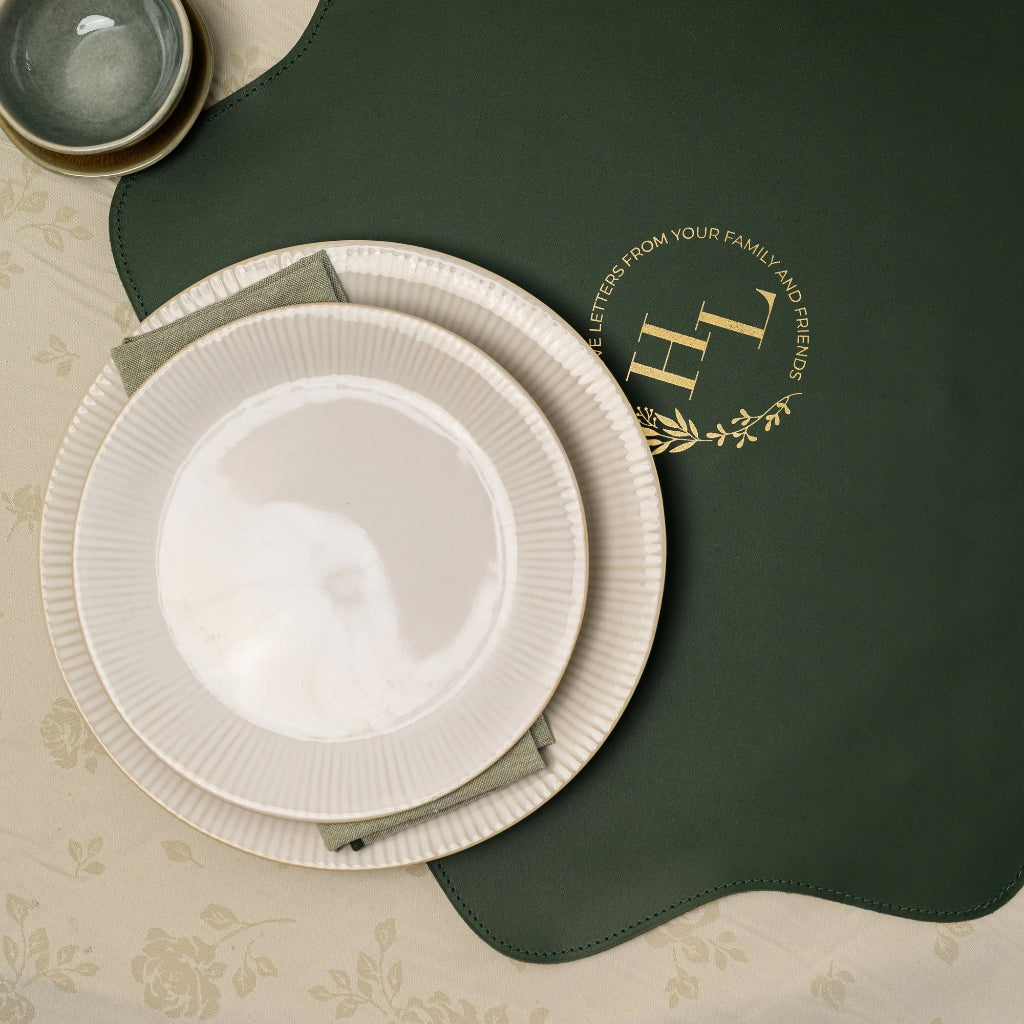 Top-down view of a unique, scalloped-edge forest green faux leather placemat with personalized gold foil branding, featuring stacked cream ribbed plates and green napkins on a floral tablecloth