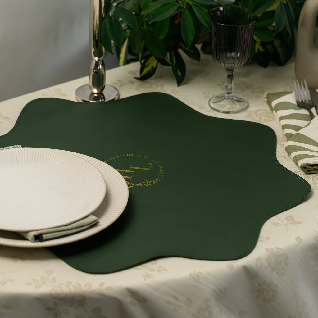 Lifestyle table setting featuring a unique scalloped-edge forest green leather placemat, styled with white ribbed plates, a wine glass, and a modern vase on a floral tablecloth
