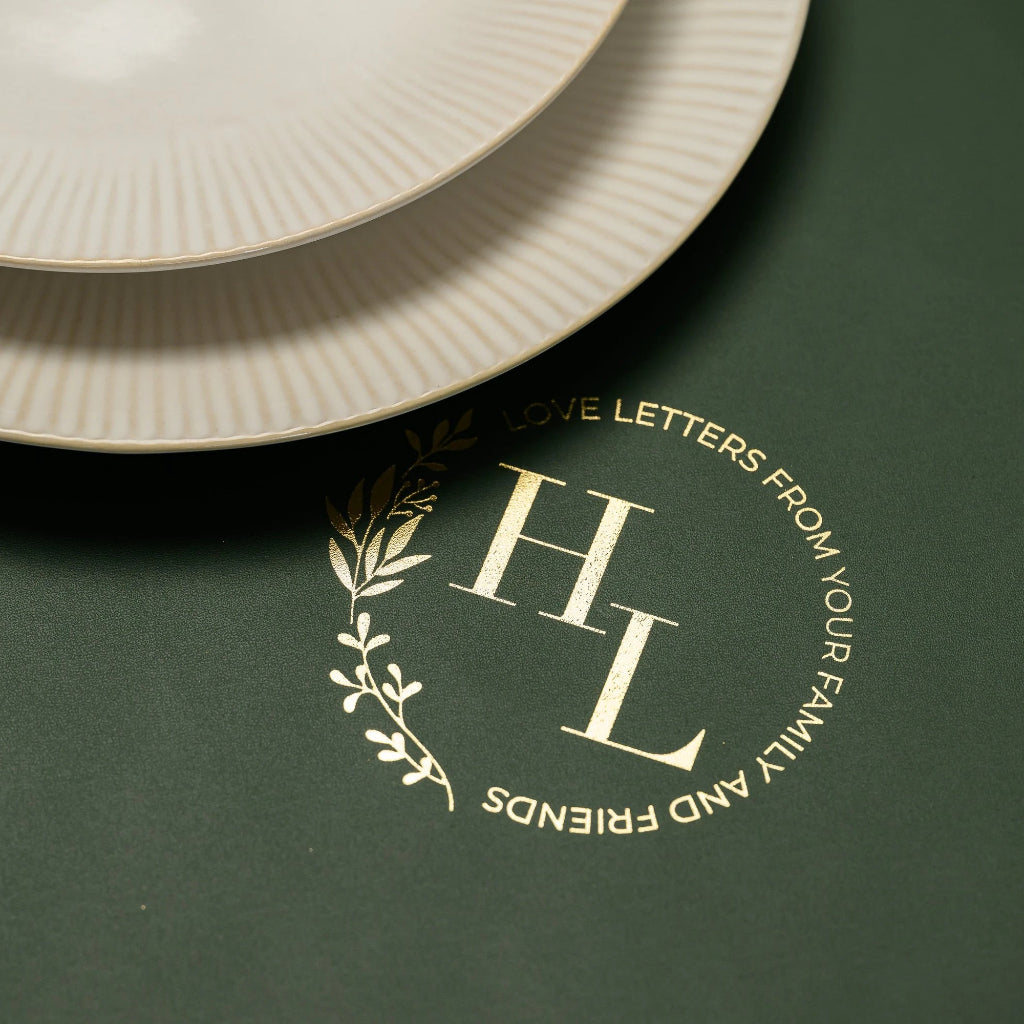Close-up of a personalized gold foil monogram 'HL' on a forest green leather placemat, featuring the circular inscription 'LOVE LETTERS FROM YOUR FAMILY AND FRIENDS' with a delicate leaf branch design