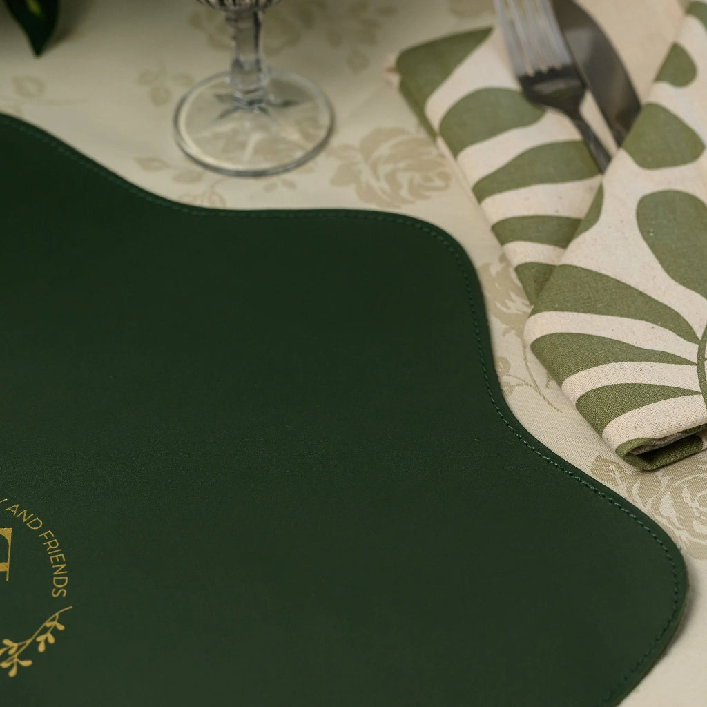 Full lifestyle shot of a unique scalloped-edge forest green leather placemat on a damask tablecloth, styled with elegant ceramic dinnerware, a wine glass, and botanical textiles