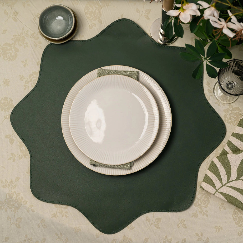 A top-down view of an elegant table setting featuring a dark forest green, scalloped-edge leather placemat as the central focus. On top of the placemat sits a stack of two cream-colored ribbed ceramic plates. The setting is arranged on a light beige floral patterned tablecloth, accompanied by a patterned green and white leaf-print napkin with cutlery, a small bowl, and decorative greenery