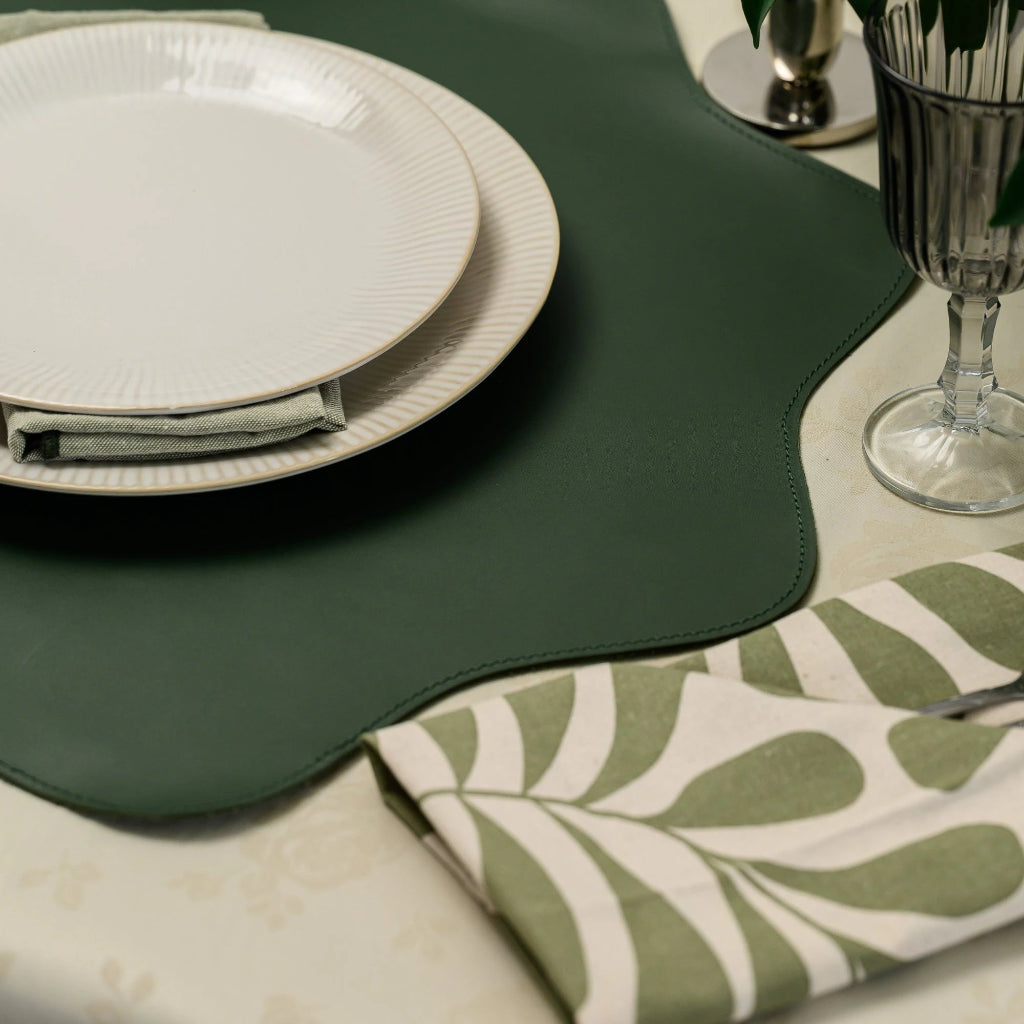 A close-up, detailed shot of a dark green leather placemat with decorative stitching and a scalloped edge. The placemat is the central element, partially covered by white ribbed ceramic plates. The smooth matte texture of the green leather contrasts with the cream floral tablecloth and a patterned leaf-print napkin nearby. A glass and green foliage are visible in the soft-focus background, emphasizing the placemat's premium look and design.