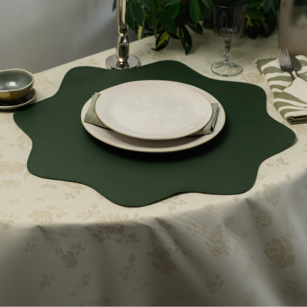 An angled side view of a table setting showcasing a dark green leather placemat with a unique wavy, scalloped border. The placemat serves as the base for a stacked pair of cream-colored ribbed plates and a folded green napkin. The setup is placed on a cream floral tablecloth, with a glass, a small bowl, and decorative foliage visible in the blurred background, highlighting the placemat's texture and shape